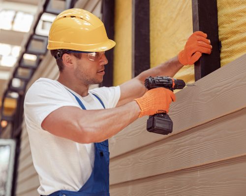 Handsome young man construction worker wearing safety helmet and work gloves while installing exterior wood siding