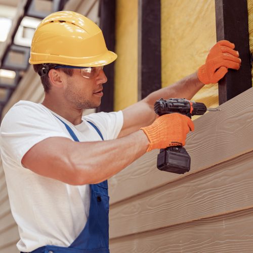 Handsome young man construction worker wearing safety helmet and work gloves while installing exterior wood siding